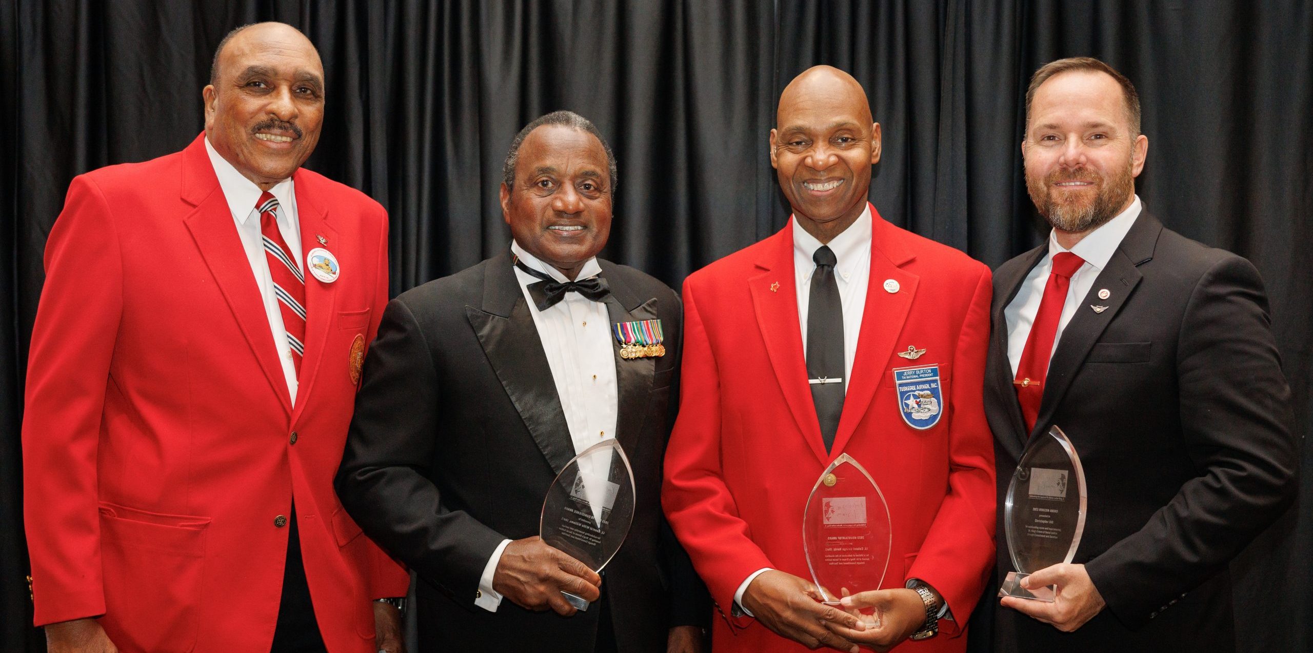 Group of four men standing in line for a picture. Three of them are holding awards in their hands