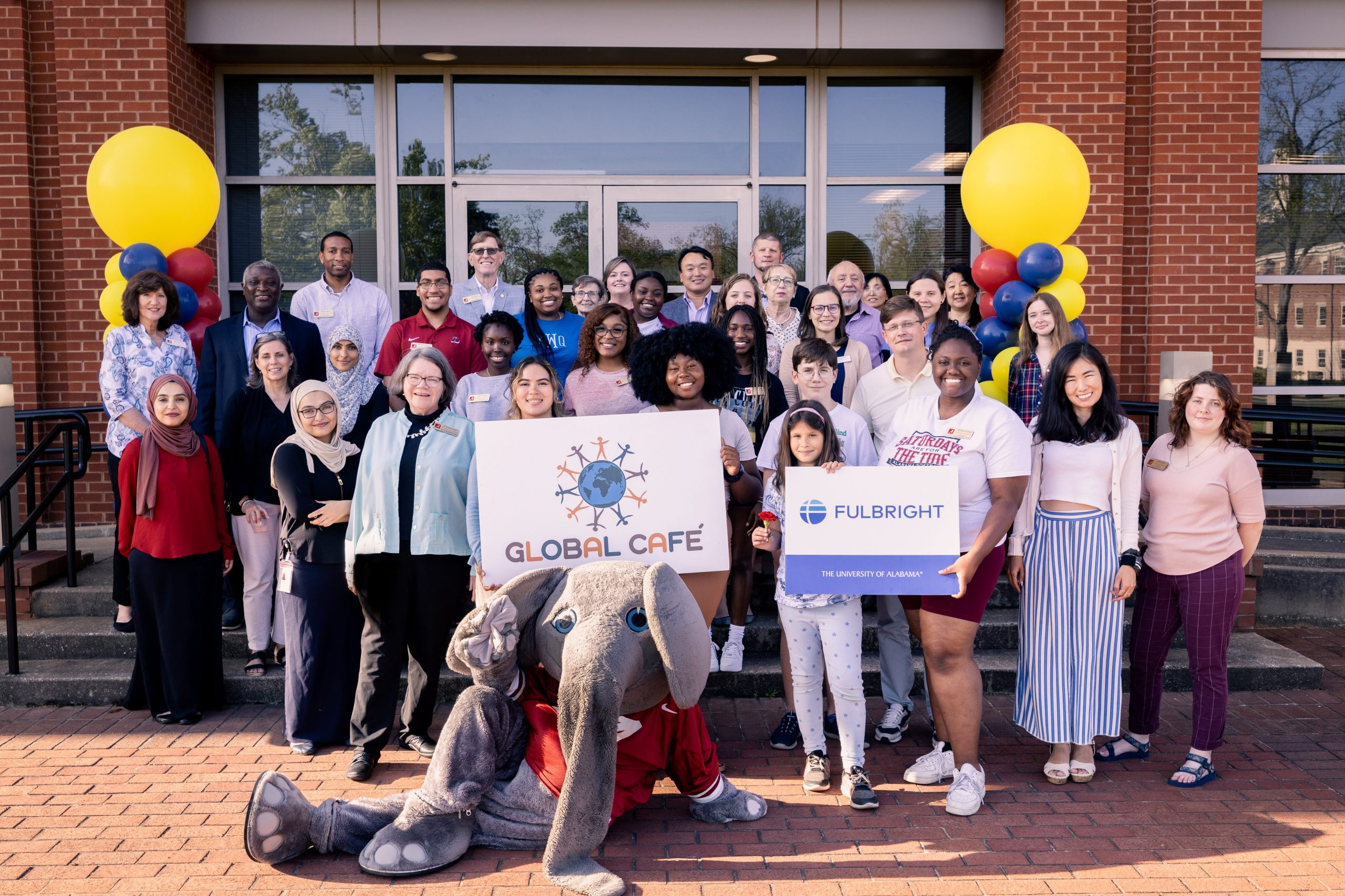 Group of Global Café volunteers, language partners, former Fulbright recipients and members of the University community posing with Big Al, yellow balloons, and Global Cafe logos