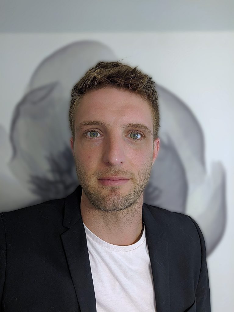 Headshot of Mathew Cieśla wearing a black blazer and white shirt in front of a flower wallpaper