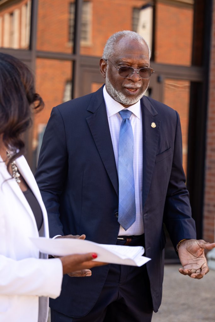 General Dr. David Satcher wearing a dark suit and blue tie talking to a woman while standing in front of a bunch of windows outside