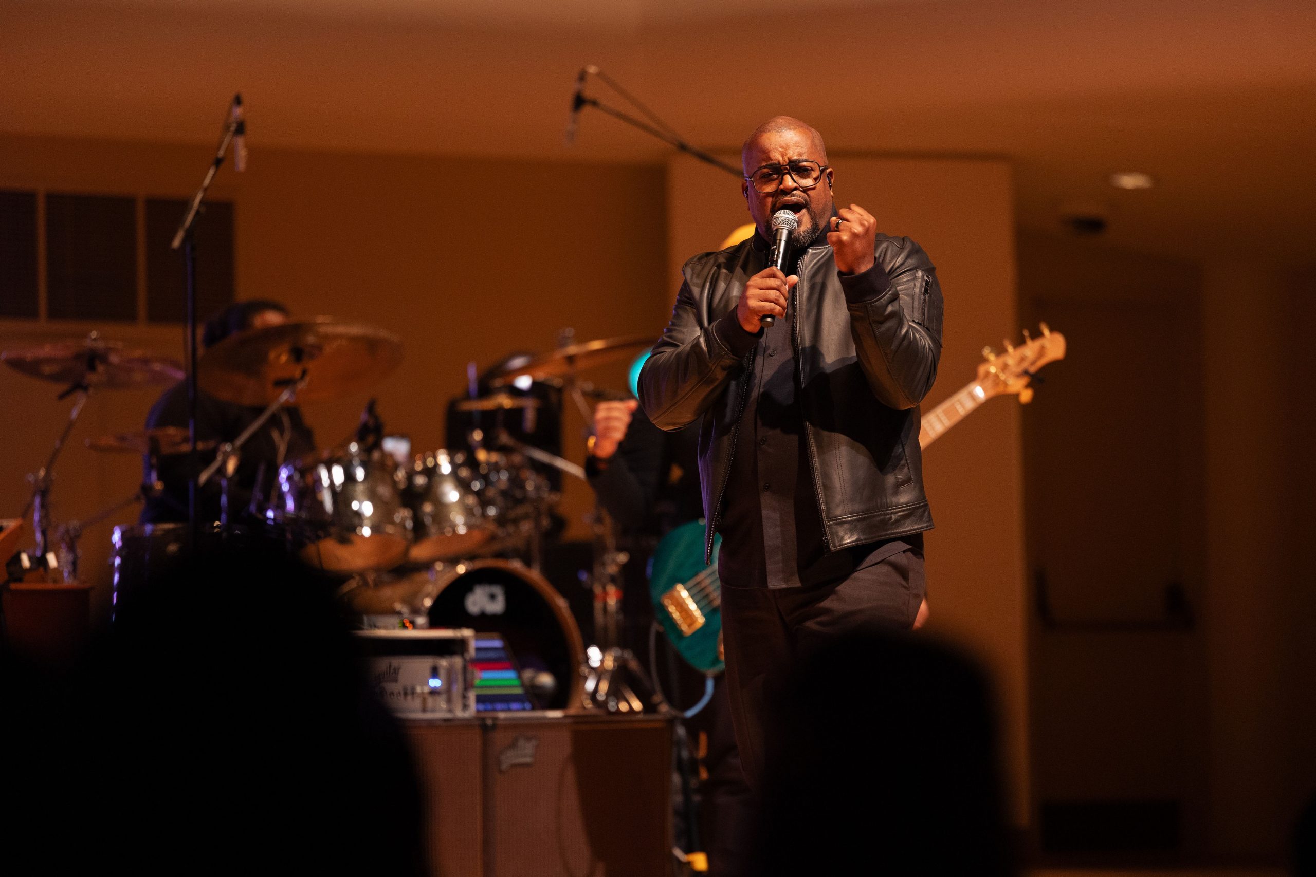 Man dressed in a black leather jack singing with a drummer, a guitarist behind him