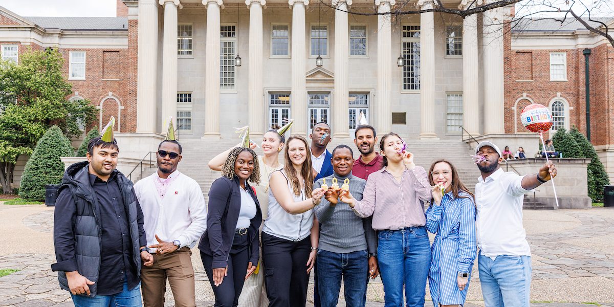 A group of people wearing party hats and holding party props pose in front of a campus building