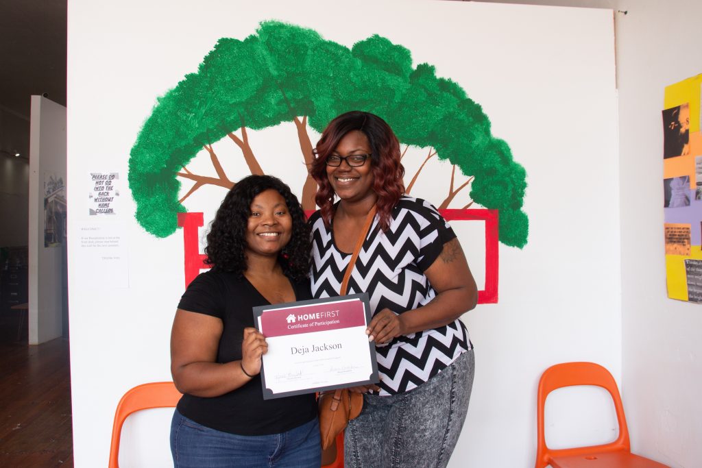 Two people standing in front of a mural of a tree holding a certificate