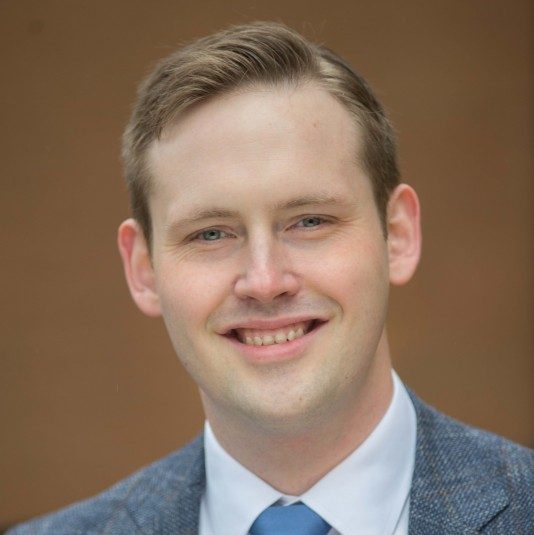 Headshot of Nick Beadle wearing a plaid suit and blue tie