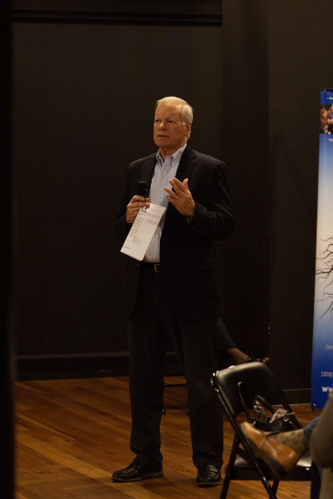 Older man in a suit speaking into a microphone will holding a piece of paper