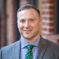 Headshot Jordan Carpenter wearing a gray suit jacket, blue paid shirt, and green patterned tie